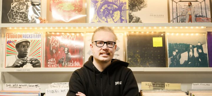 Henkilöllä on musta huppari ja silmälasit. Taustalla hylly värikkäitä LP-levyjä.